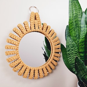 May include: A round mirror with a woven, mustard yellow frame. The frame is made of thick rope and has a sunburst design. The mirror is hanging on a white wall.