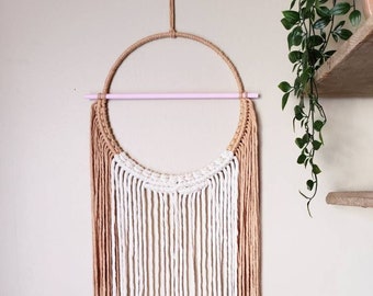 May include: A macrame wall hanging featuring a circular design with a natural rope frame. The piece incorporates white, tan, and light pink cords, creating a layered, textured aesthetic. The wall decor is suspended from a natural rope.