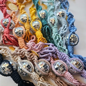 May include: A collection of macrame hanging ornaments in various colors, each featuring a small spherical disco ball with mirrored facets. The cotton cord is knotted in a spiral pattern, ending in a fringe, with a small circular tag attached.