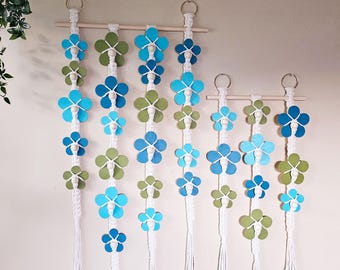 May include: A set of four macrame wall hangings with wooden dowels and blue and green felt flower shapes. The hangings are different sizes and are arranged in a row on a white wall.
