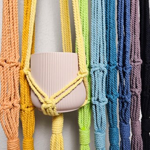 May include: A macrame plant hanger in a rainbow of colors, including peach, yellow, green, blue, purple, and black. The plant hanger holds a light pink, ribbed ceramic pot. The rope is knotted to create a decorative and functional design.