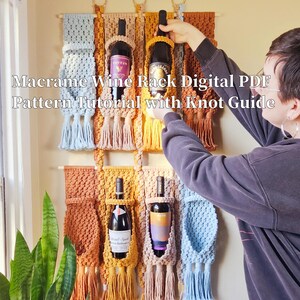 May include: Macrame wine rack with four bottle holders. The rack is made with natural colored cord and has a light blue, tan, and mustard yellow color scheme. The rack is hanging on a wall with four bottles of wine in the holders.