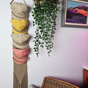 May include: A macrame hat organizer in beige holds six baseball caps in various colors: tan, yellow, and pink. The organizer hangs from a rope and has a fringed bottom. A hanging plant and framed landscape painting are in the background.