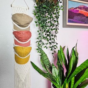 May include: A macrame hat holder with four baseball caps in tan, coral, and yellow. The caps are displayed on a white wall, with a hanging plant and a framed landscape painting in the background. The macrame holder is made of cream-colored rope.