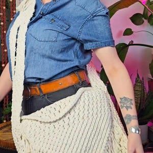May include: Cream-colored macrame shoulder bag with a fringed bottom. The bag is worn over a denim shirt and black jeans. The bag has a woven texture and a long strap.