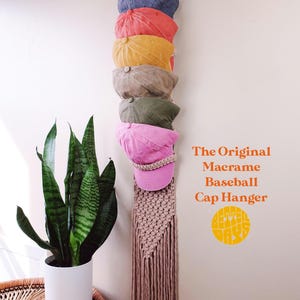 Puede incluir: Un colgador de gorras de béisbol de macramé en color beige, con un aro superior circular y una base de flecos anudados. Sostiene seis gorras de béisbol en azul, coral, amarillo, beige, verde oliva y rosa. El texto dice: The Original Macrame Baseball Cap Hanger.