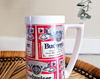 Könnte beinhalten: Ein weißer Bierkrug aus Kunststoff mit einem sich wiederholenden rot-weiß-blauen Budweiser-Etikettendesign. Der Krug hat einen zylindrischen Körper mit einem weißen Henkel und einem weißen Rand. Der Aufdruck zeigt das Budweiser-Logo, Text und Wappen.