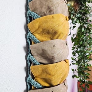 May include: A wall-mounted hat organizer with five baseball caps in shades of tan and yellow. The caps are suspended from a teal macrame hanger. The caps are arranged vertically, showcasing their curved brims and top buttons. The background is a white wall.