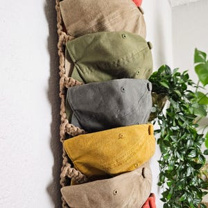 May include: A macrame hat holder displaying seven baseball caps in various colors: red, tan, olive green, gray, yellow, and two more tan and red caps. The caps are made of a soft fabric and have a curved brim. The holder is made of woven rope.