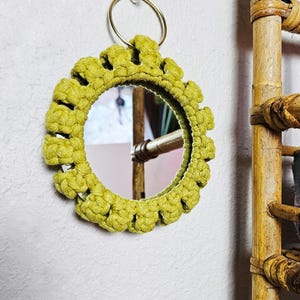 May include: A circular wall mirror framed by a thick, textured border of knotted olive green cord. The mirror features a simple gold-toned metal ring at the top for hanging, with the knotted cord creating a scalloped, petal-like edge around the glass.