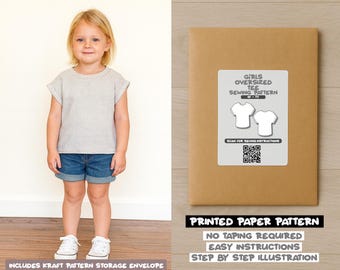 Op de afbeelding: Een jong meisje draagt een grijs t-shirt en een spijkerbroekshort, naast een bruine papieren patroon-envelop. De envelop heeft een label met de tekst "Girls Oversized Tee Sewing Pattern" en een QR-code. De tekst "Printed Paper Pattern" is ook zichtbaar.