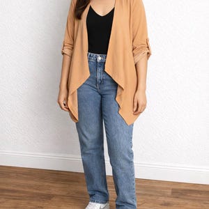 Woman wearing a tan Waterfall Jacket, black top, blue jeans, and white sneakers against a white wall.