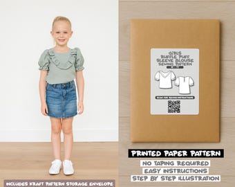 Op de afbeelding: Een jong meisje draagt een saliegroene blouse met pofmouwen en een spijkerbroekrok. Naast haar staat een bruine papieren envelop met het label "GIRLS RUFFLE PUFF SLEEVE BLOUSE SEWING PATTERN". De envelop vermeldt ook "PRINTED PAPER PATTERN".