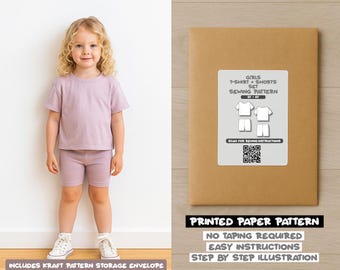 Op de afbeelding: Een jong kind draagt een lila t-shirt en short set, naast een bruine papieren envelop. De envelop heeft een wit label met een naaipatroondiagram en de tekst "GIRLS T-SHIRT + SHORTS SET SEWING PATTERN". De tekst "PRINTED PAPER PATTERN" is ook zichtbaar.