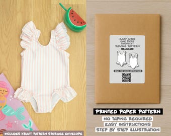 Op de afbeelding: Een baby meisje badpak met roze en witte strepen en ruches. Een watermeloen speelgoed en een naaipatroon envelop zijn ook zichtbaar. Het patroon bevat eenvoudige instructies.