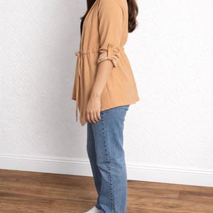 Side view of woman wearing a tan Waterfall Jacket, black top, blue jeans, and white sneakers against a white wall.