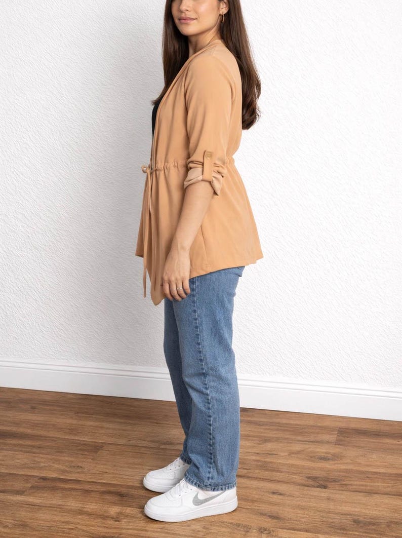 Side view of woman wearing a tan Waterfall Jacket, black top, blue jeans, and white sneakers against a white wall.