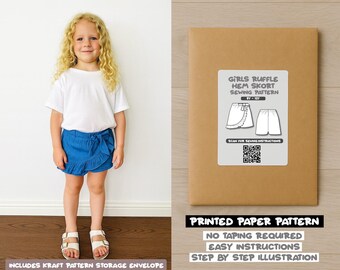 Op de afbeelding: Een jong meisje in een wit t-shirt en een blauwe ruffle short staat naast een bruine papieren patroon envelop. De envelop heeft een label met "GIRLS RUFFLE HEM SKORT SEWING PATTERN" en een QR-code. De tekst "PRINTED PAPER PATTERN" is ook zichtbaar.