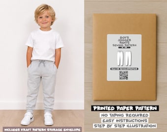 May include: A young boy wearing a white t-shirt and gray sweatpants stands next to a brown paper pattern envelope. The envelope has a white label with the text "BOYS JOGGER PANTS SEWING PATTERN" and a QR code. The text "PRINTED PAPER PATTERN" is also visible.