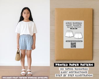 Op de afbeelding: Een jong persoon draagt een wit t-shirt en een blauwe korte broek met bloemenpatroon, met een kleine geweven tas. Daarnaast een bruine papieren envelop met het label "GIRLS SHIRRED WAIST SHORTS SEWING PATTERN". De tekst "PRINTED PAPER PATTERN" is ook zichtbaar.