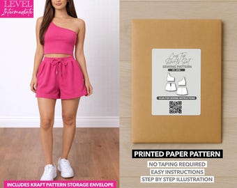 Può includere: Una donna indossa un completo crop top e pantaloncini rosa fucsia. L'outfit comprende un top monospalla e pantaloncini con coulisse. A destra, una busta di carta kraft con un'etichetta per il modello di cucito. L'etichetta recita "Crop Top Shorts Set Sewing Pattern".