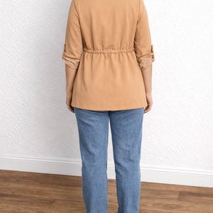 Back view of woman wearing a tan Waterfall Jacket, blue jeans, and white sneakers against a white wall.