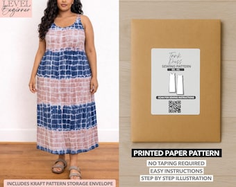 May include: A woman wearing a tie-dye tank dress in shades of blue and pink. The dress is paired with sandals. Next to the woman is a brown paper pattern envelope with a white label that reads "Tank Dress Sewing Pattern". The envelope also says "Printed Paper Pattern".