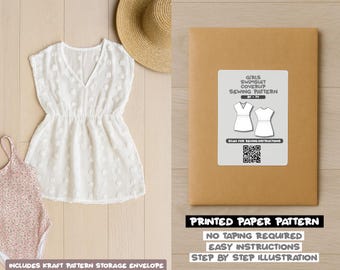 Op de afbeelding: Een witte cover-up jurk met een V-hals en een strohoed worden gepresenteerd op een licht houten oppervlak. Een kraftpapieren envelop met een naaipatroonlabel is ook aanwezig. Het label luidt "Girls Swimsuit Coverup Sewing Pattern".