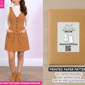 Op de afbeelding: Een vrouw draagt een bruine corduroy overall mini-jurk met een witte coltrui. De jurk heeft grote zakken en knoopdetails. Een kraftpapieren envelop met een naaipatroonlabel staat rechts. Het label luidt "Overall Mini Dress Sewing Pattern".