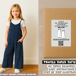 May include: A young girl in a navy jumpsuit with white polka dots, a white t-shirt, and white trainers. The jumpsuit features tie shoulder straps and pockets. Beside her is a brown paper pattern envelope with text: "Girls Tie Shoulder Jumpsuit Sewing Pattern."
