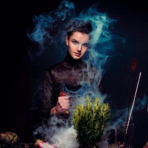 May include: A woman in a black sheer top stands in a cloud of blue smoke, holding a glass with a red liquid. A green plant sits in front of her, and there are various glass bottles and other objects on the table.