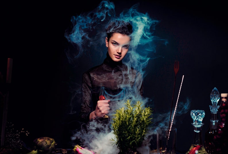 May include: A woman in a black sheer top stands in a cloud of blue smoke, holding a glass with a red liquid. A green plant sits in front of her, and there are various glass bottles and other objects on the table.