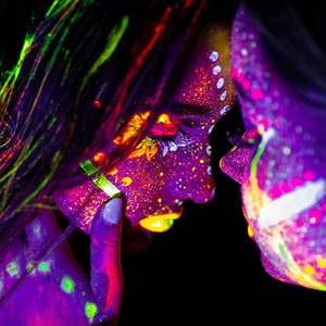 May include: Two people with neon paint on their faces and bodies, looking at each other. The paint is in shades of purple, green, yellow, and orange.