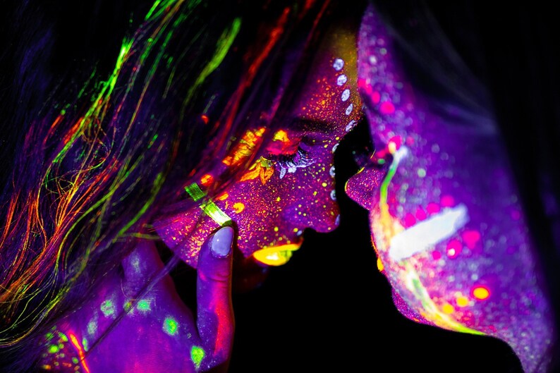 May include: Two people with neon paint on their faces and bodies, looking at each other. The paint is in shades of purple, green, yellow, and orange.
