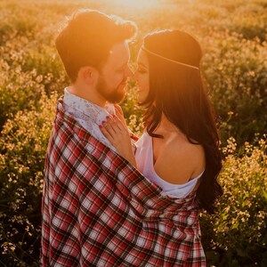 May include: A couple standing in a field of wildflowers, embracing and looking at each other. The man is wearing a plaid shirt and the woman is wearing a white top. The sun is setting behind them, casting a warm glow on the scene.