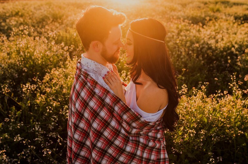 May include: A couple standing in a field of wildflowers, embracing and looking at each other. The man is wearing a plaid shirt and the woman is wearing a white top. The sun is setting behind them, casting a warm glow on the scene.