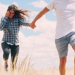 May include: A couple holding hands and running through a field of tall grass. The woman is wearing a plaid shirt and black pants. The man is wearing a white t-shirt and blue shorts.