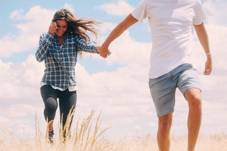 May include: A couple holding hands and running through a field of tall grass. The woman is wearing a plaid shirt and black pants. The man is wearing a white t-shirt and blue shorts.