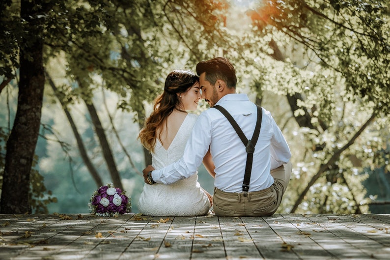 May include: A couple in wedding attire, sitting on a wooden deck, with a bouquet of purple and white flowers in front of them. They are looking at each other and smiling.