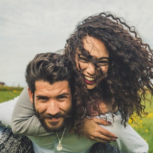 May include: A couple is laughing and having fun outdoors. The woman is wearing a white shirt and has long curly hair. The man is wearing a gray shirt and has a beard. They are in a field of green grass and yellow flowers.