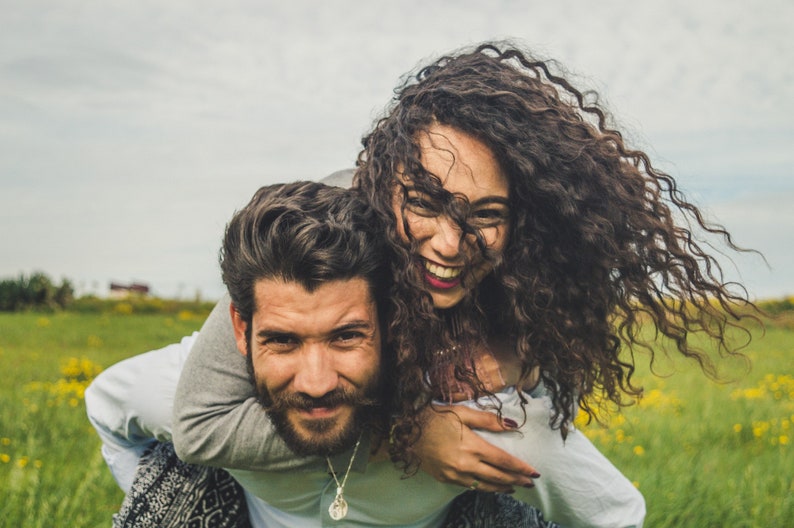 May include: A couple is laughing and having fun outdoors. The woman is wearing a white shirt and has long curly hair. The man is wearing a gray shirt and has a beard. They are in a field of green grass and yellow flowers.