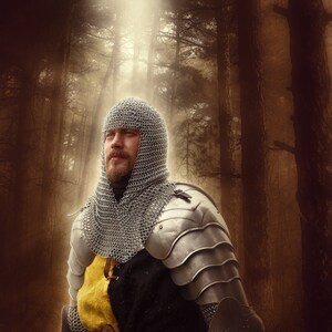 May include: A knight in chainmail armor stands in a forest. He is wearing a helmet and has a beard. The forest is dark and mysterious, with tall trees and a light shining through the branches.