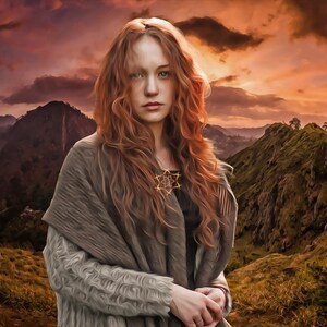 May include: A woman with long, curly red hair stands in front of a mountain range at sunset. She is wearing a gray knitted shawl and a necklace with a star-shaped pendant.