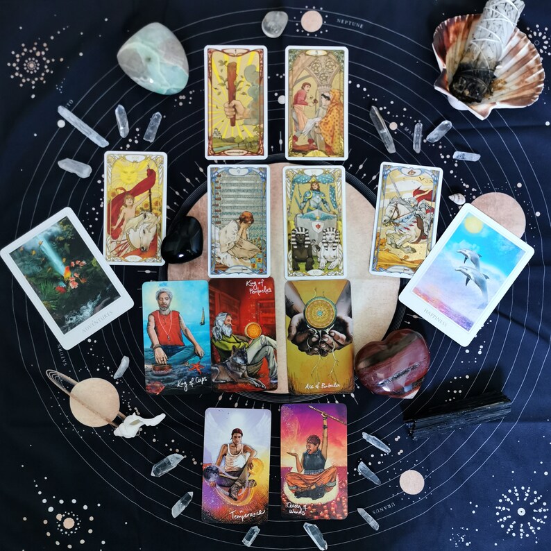 May include: A tarot card spread on a black background with a variety of crystals and other objects. The cards are arranged in a circle with the "King of Cups" card in the centre. The cards are from the "Wild Unknown Tarot" deck.