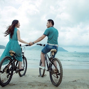 May include: A couple riding bicycles on a sandy beach with a mountain range in the background. The woman is wearing a light blue dress and the man is wearing a teal shirt and blue jeans.