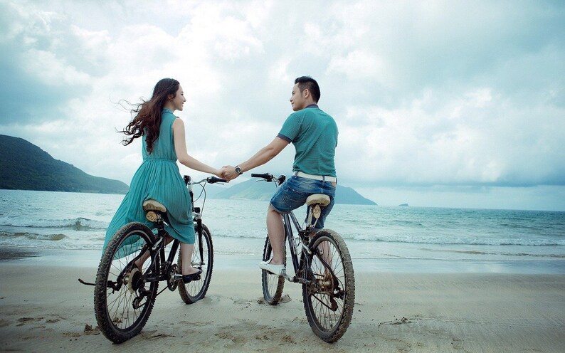 May include: A couple riding bicycles on a sandy beach with a mountain range in the background. The woman is wearing a light blue dress and the man is wearing a teal shirt and blue jeans.