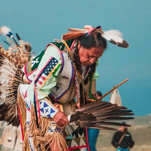 May include: A Native American man in traditional regalia, wearing a headdress with feathers, a beaded vest, and a fringed skirt, performs a dance with a feathered fan.