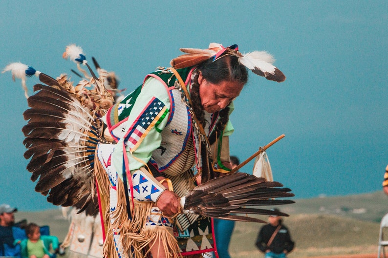May include: A Native American man in traditional regalia, wearing a headdress with feathers, a beaded vest, and a fringed skirt, performs a dance with a feathered fan.