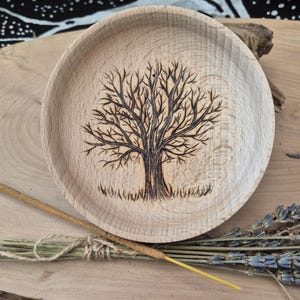 May include: A wooden incense holder with a tree design burned into the surface. The tree is dark brown and has detailed branches. The holder is surrounded by dried lavender, an incense stick, and other natural elements.