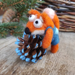 May include: A felt fox toy with orange fur, white accents, and blue overalls, holding a blue and brown pine cone.